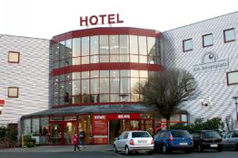 Gebäude mit Aufschrift "Hotel" und "Rewe", moderne Architektur, parkende Autos vorne.