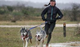 Mujer corre en un camino lluvioso acompañada por dos perros husky atados con correas.