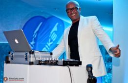 Ein DJ mit weißem Sakko lächelt hinter einem Mischpult mit Laptop und Mikrofon.