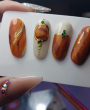 Uñas decoradas en tonos marrón y oro con detalles de pedrería y esmalte brillante.
