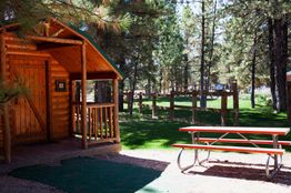 Log cabin with porch beside a red picnic table in a forested park area.