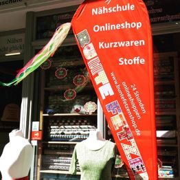 Rotes Banner vor Stoffladen mit der Aufschrift "Nähschule, Onlineshop, Kurzwaren, Stoffe."