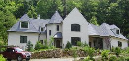 A large white house with a slate roof and red car, surrounded by greenery and trees.