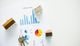 Charts, house models, and keys on a desk symbolize real estate and financial analysis.