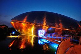 Futuristisches Gebäude bei Nacht, beleuchtet in Blau und Orange, mit Spiegelung im Wasser.