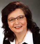 Eine lächelnde Frau mit Brille und dunkelbraunem Haar vor grauem Hintergrund.