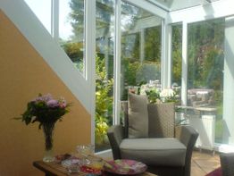 Wintergarten mit Korbstuhl, Blumenstrauß und Tisch, durchsonnt von Tageslicht.