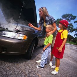 Woman and two kids look at a steaming car engine on the roadside under a clear sky.