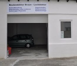 Autowerkstatt mit offenen Tor, grünes Auto innen, Schild mit Werkstattservice-Details oben.