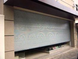 Loja fechada com porta metálica de enrolar cinza, fachada de concreto, e vitrines inferiores.