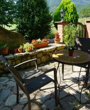 Patio con sillas, mesa y macetas, rodeado de vegetación con montañas al fondo en un día soleado.