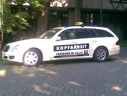 Weißes Auto mit "Kopfarbeit Friseure in Köln"-Werbung steht auf gepflastertem Parkplatz.