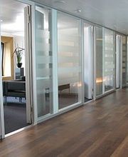 Büroflur mit Glaswänden, Holzboden und Blick in ein Zimmer mit Sesseln und Pflanze.