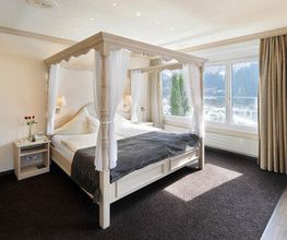 Helles Schlafzimmer mit Himmelbett, Vorhängen und Panoramafenster, Blick auf Berglandschaft.