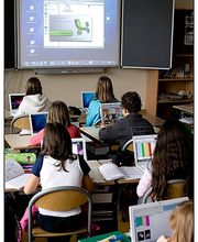 Schulkinder lernen mit Laptops in einem Klassenzimmer; eine Präsentation wird projiziert.