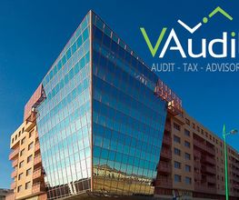 Edificio moderno con fachada de vidrio, logo de VAudit y lista de servicios en esquina superior derecha.