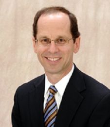 Smiling man in glasses wearing a suit and striped tie against a beige background.