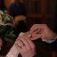 Zwei Personen tauschen Ringe bei einer Hochzeit. Ein Brautstrauß ist im Hintergrund sichtbar.
