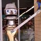 Robot sculpture made of metal kitchenware with a quirky face, standing by a glass door.