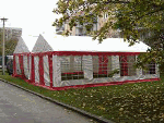 Ein rot-weißes Festzelt steht auf einer Wiese, umgeben von Bäumen und Gebäuden.