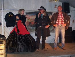 Drei Personen in Westernkleidung auf einer Bühne, eine Frau im Kleid und zwei Männer in Cowboy-Outfits.