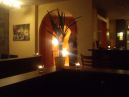 Verschwommener Raum mit Kerzen, Pflanze in der Mitte, Person im Hintergrund bei gedämpftem Licht.