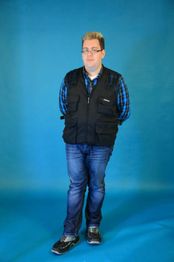 Person in kariertem Hemd und Weste steht vor blauem Hintergrund, die Hände hinter dem Rücken.