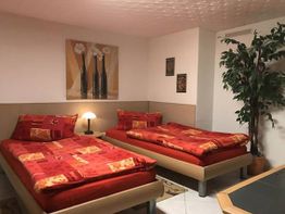 Zwei Einzelbetten mit roten Bettdecken, Lampe dazwischen, Bilder und Pflanze im Raum.