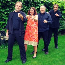 Vier Personen posieren lachend im Garten auf dem Rasen vor einer Hecke und zeigen in die Kamera.