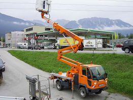 Ein orangefarbener Hebebühnen-LKW bei einer Straße, mit Bergen im Hintergrund.