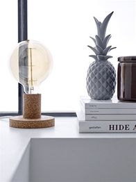 Lampe mit Korkfuß, graue Ananas-Deko und gestapelte Bücher auf hellem Tisch vor Fenster.