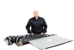 Mann in schwarzem Hemd mit DJ-Equipment: Laptop, Kopfhörer und Mikrofon vor weißem Hintergrund.