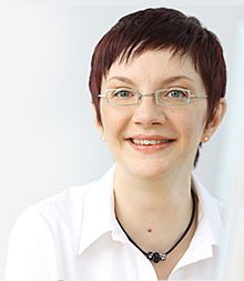 Eine lächelnde Person mit kurzen Haaren, Brille und weißem Hemd vor hellem Hintergrund.