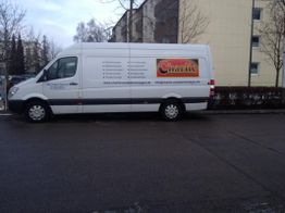Weißer Lieferwagen mit "Charlite"-Werbung, steht auf nasser Straße vor einem gelben Gebäude.