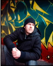 Mann in schwarzer Kleidung mit Mütze vor bunter Graffiti-Wand, schaut nach links.