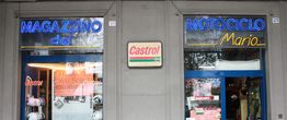 Due vetrine di negozi con insegne luminose: "Magazzino del Motociclo" e logo Castrol al centro.