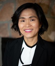 Portrait of a person smiling, wearing a black blazer and white shirt in front of a stone wall.