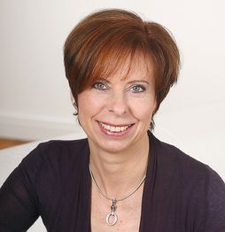 Porträt einer lächelnden Frau mit kurzen, rotbraunen Haaren und violettem Oberteil vor hellem Hintergrund.