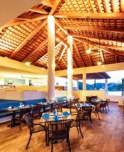 Restaurante de techo alto y madera, muebles oscuros, vista al mar y palmeras al atardecer.