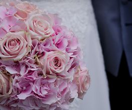 Bouquet di rose e ortensie rosa tenuto da una sposa in abito bianco e uomo in abito blu.