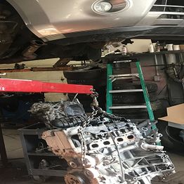 Car engine removal in a garage; vehicle elevated, engine suspended on a hoist.