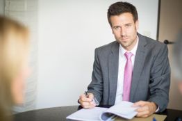 Ein Mann im Anzug sitzt am Tisch mit einem offenen Buch und schaut in die Kamera.