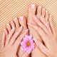 Hands and feet with French manicure, holding a pink flower on a bamboo mat background.