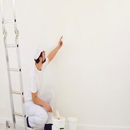 Painter in white outfit kneels, pointing at a white wall beside a ladder and paint bucket.