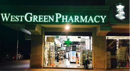 Street view of West Green Pharmacy with illuminated sign and glass entrance at night.