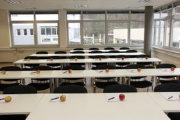 Leerer Schulungsraum mit Tischen, darauf Äpfel und Notizblöcke mit Stiften, große Fenster im Hintergrund.