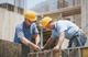 Two construction workers in hard hats secure wires at a building site.