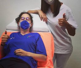 Dos personas con mascarillas, una acostada y otra de pie, mostrando pulgares arriba en señal positiva.