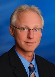 Älterer Mann mit Brille im Anzug vor blauem Hintergrund, lächelt leicht in die Kamera.