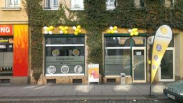 Geschäft mit gelben Luftballons und Fahne, Fenster mit Logos, Efeu an der Fassade, Straße davor.